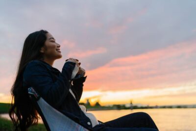 Young woman enjoying hot drink in nature during sunset by lake
